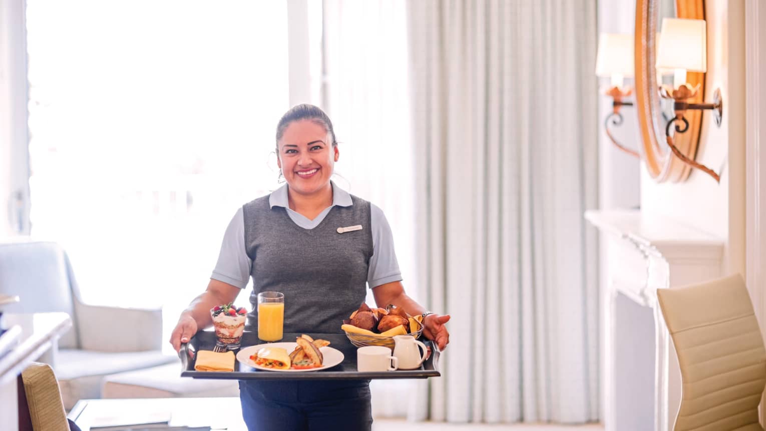 Smiling server carries in-room breakfast tray in elegant white hotel room