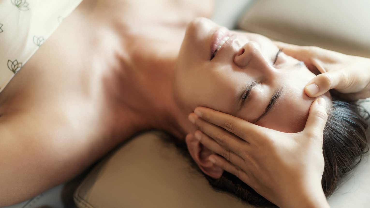 Spa facial, two hands rest on woman's forehead as she closes her eyes, lays under sheet on treatment table
