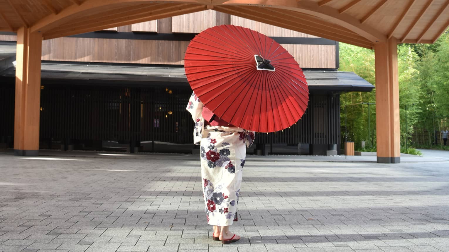 Rear view of a person under a wooden structure, in a white, flower-printed kimono, partially hidden by a red paper umbrella.