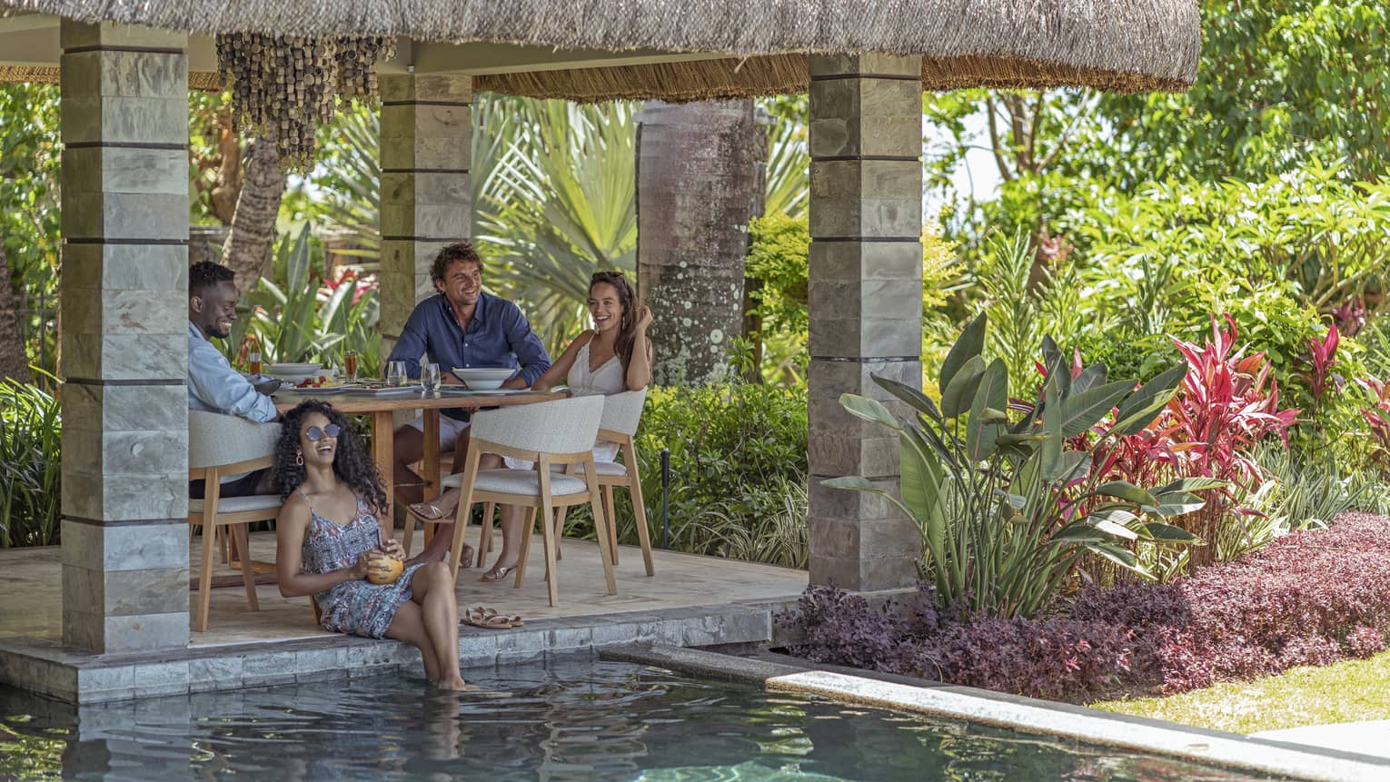 Four people sit at dining table under thatched-roof patio, next to pool