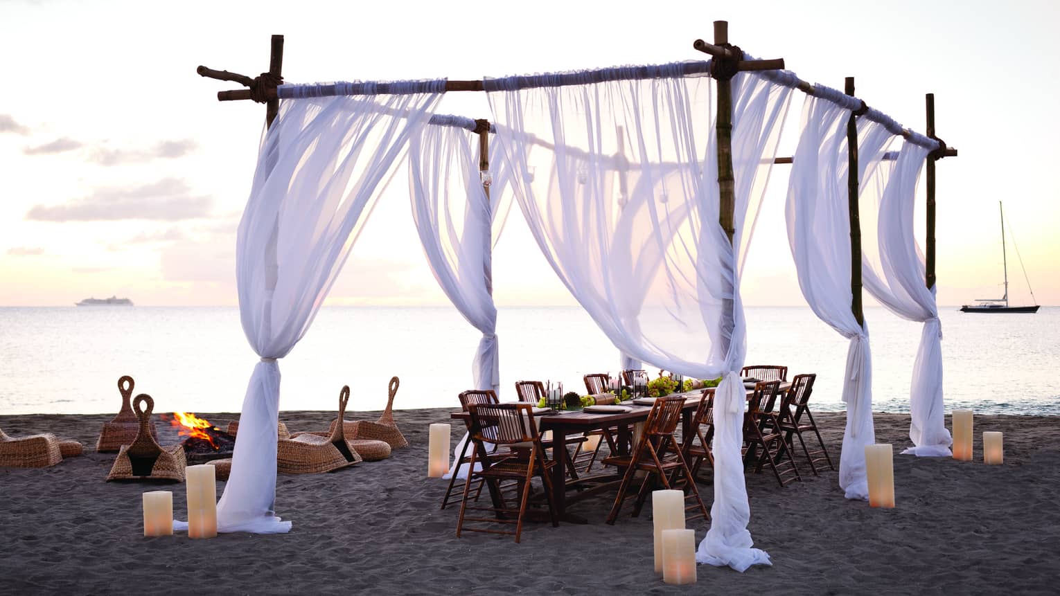 Sheer white curtains draped across wood posts around dining table on beach at sunset