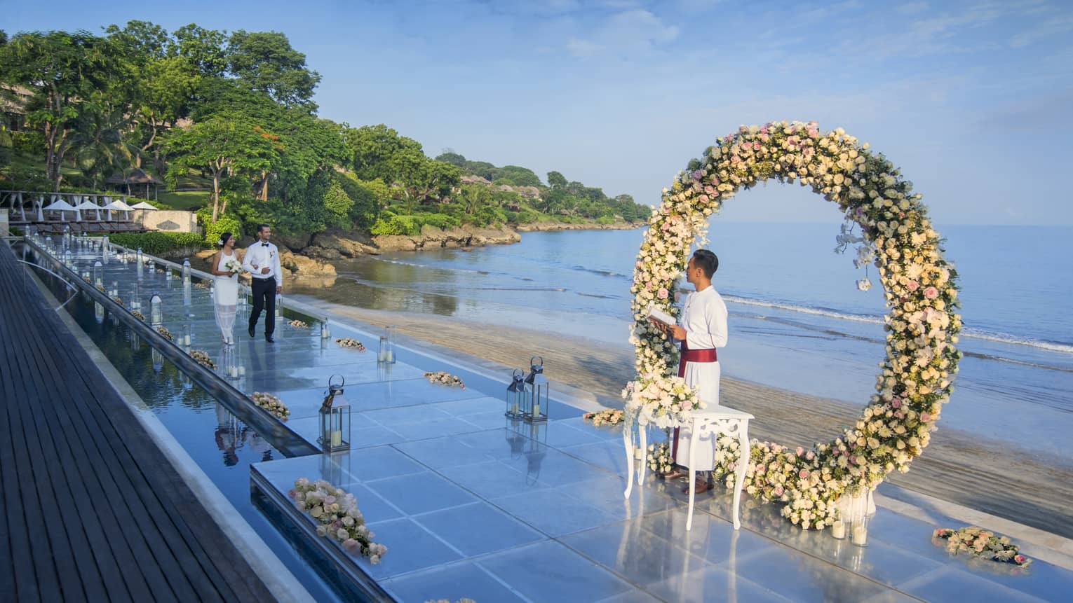 A couple walks down the aisle toward a flower arch during an outdoor wedding ceremony