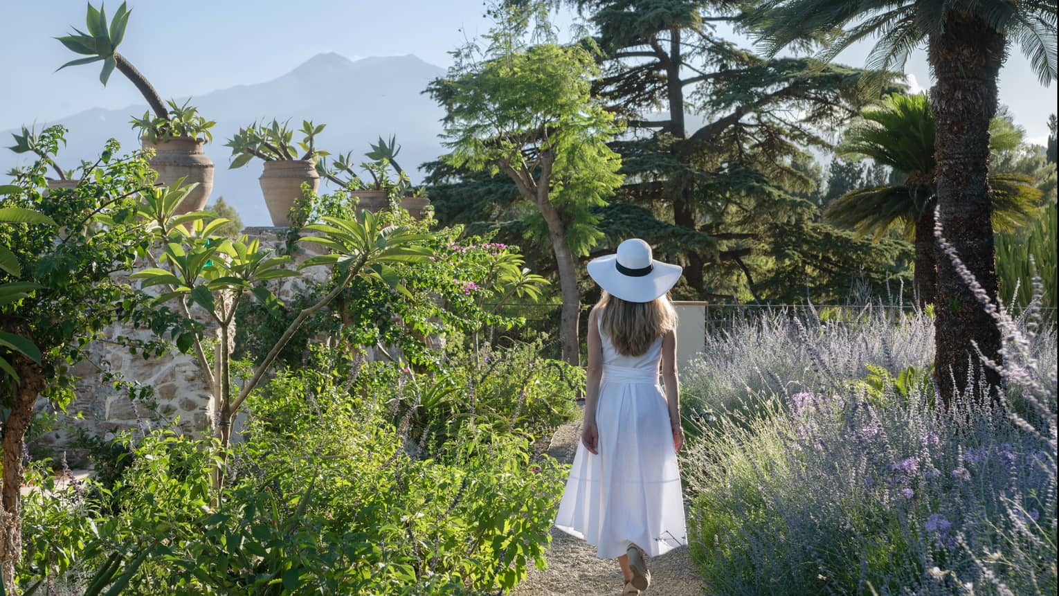 Woman in white dress walking on path in garden with Mount Etna in distance