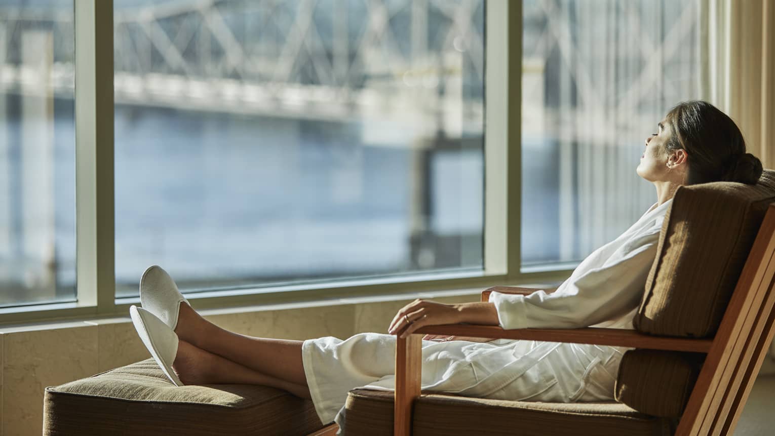 Woman wearing white spa bathrobe and slippers lounges in chair by window