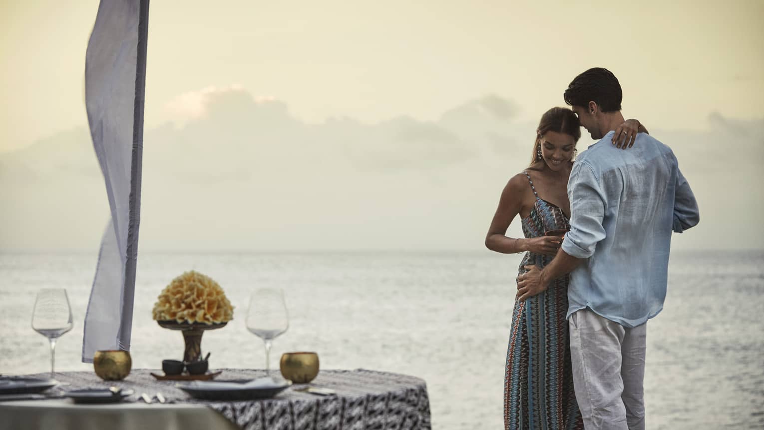 Couple embraces beside private dining table overlooking ocean at sunset