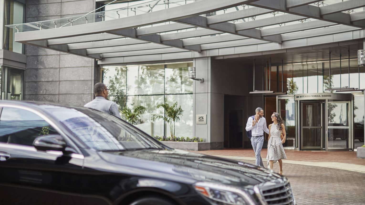 A man and woman walking towards a black sedan after leaving a hotel, with a hotel staff member standing by the car.