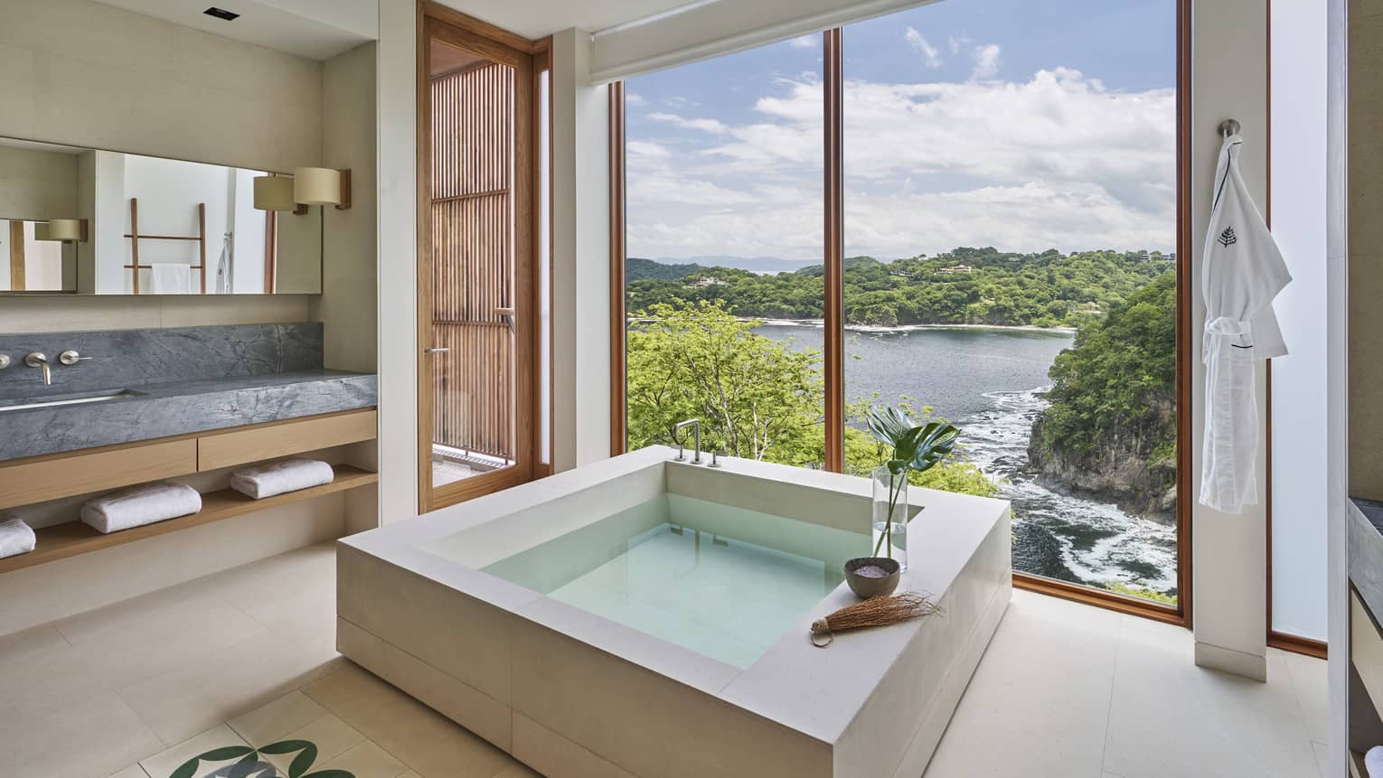 Bathroom with square stone tub; a wall of windows look out to the sea and trees