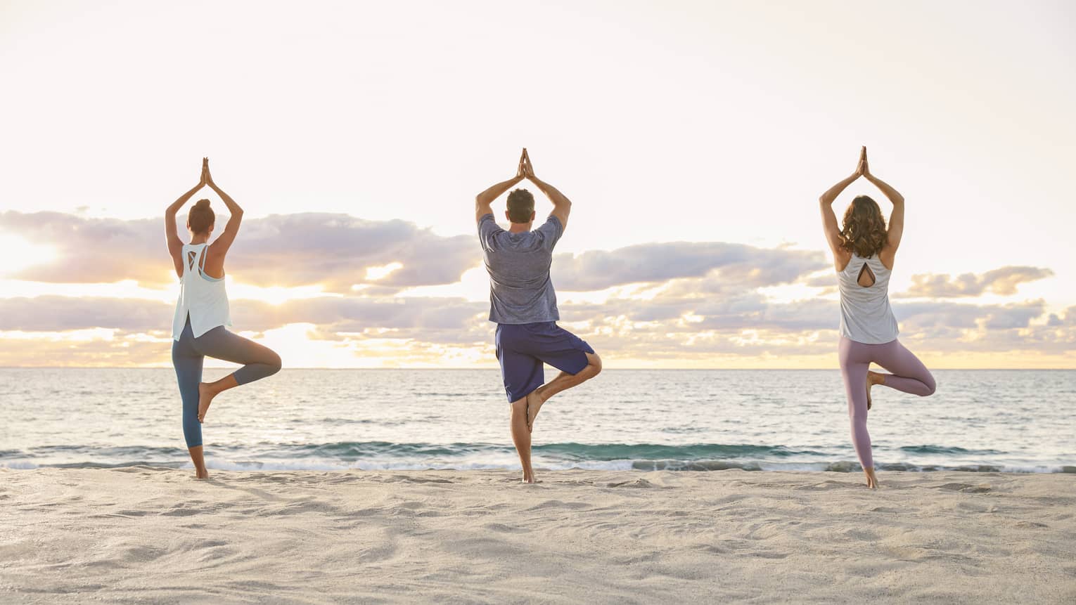 Backs of three people standing in yoga pose on sand beach at sunrise