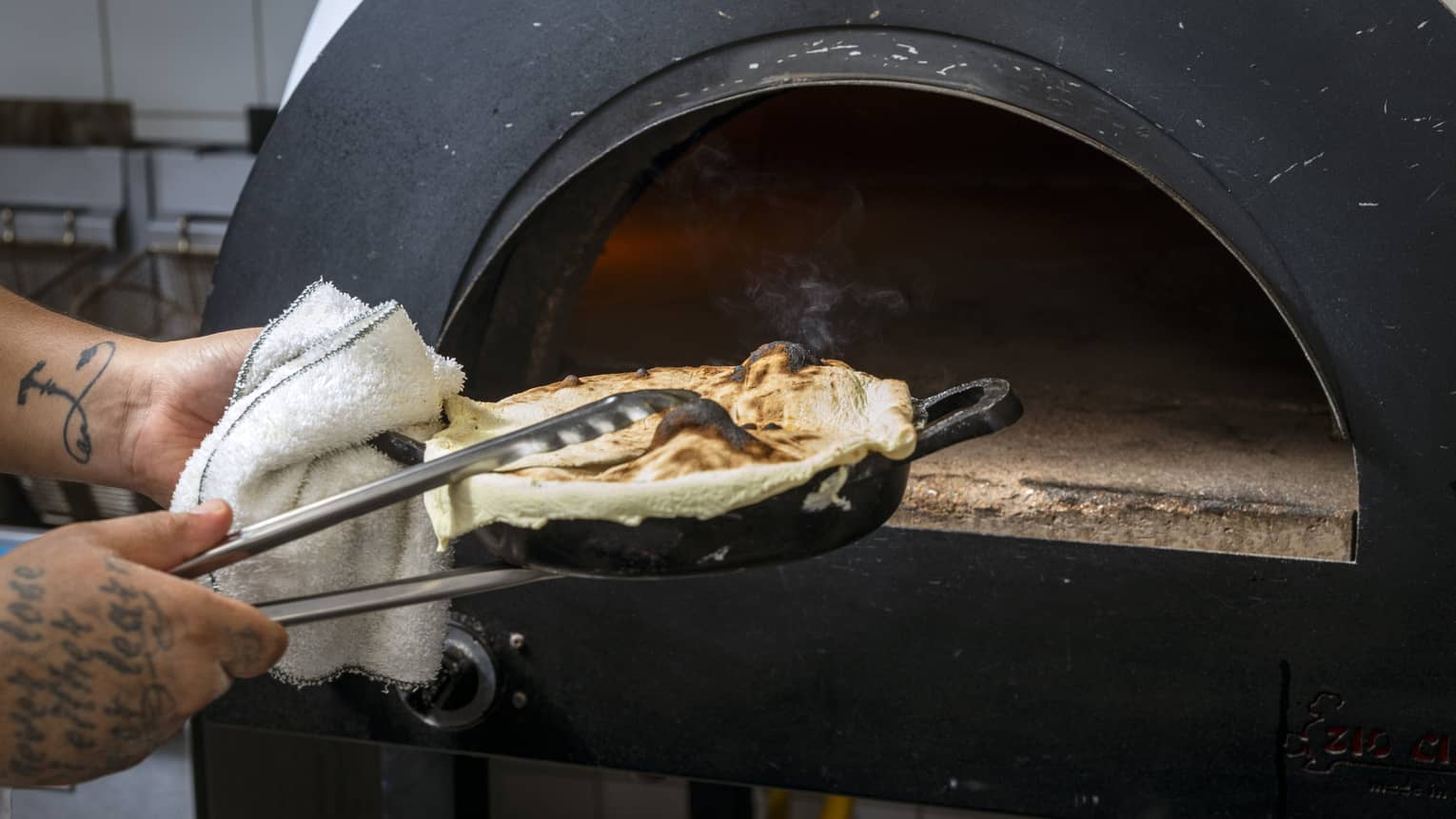 Tattooed hands pull a fresh-baked pita out of a wood oven