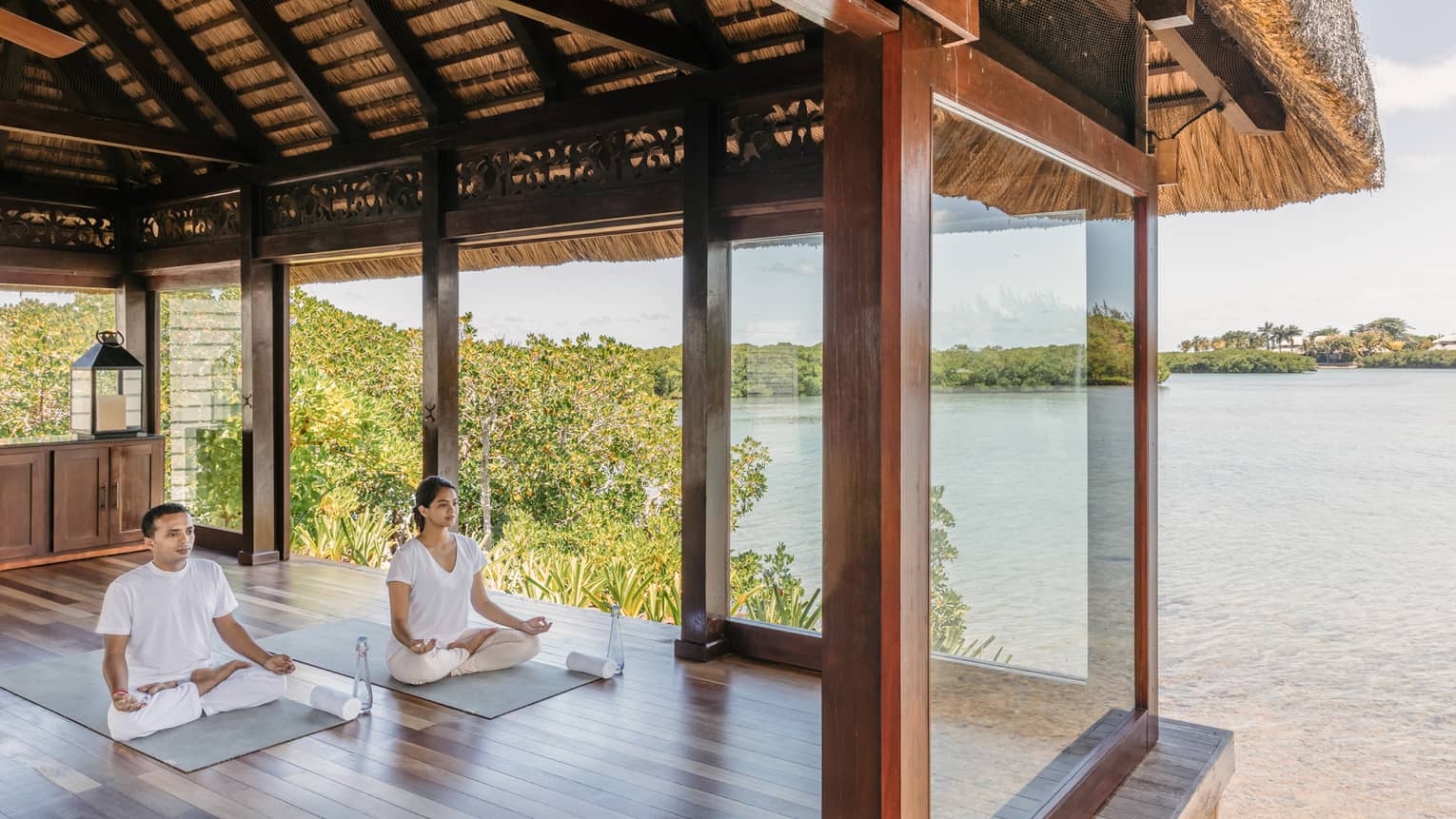 Two people wearing white seated in yoga poses in open-air spa yoga pavilion by beach