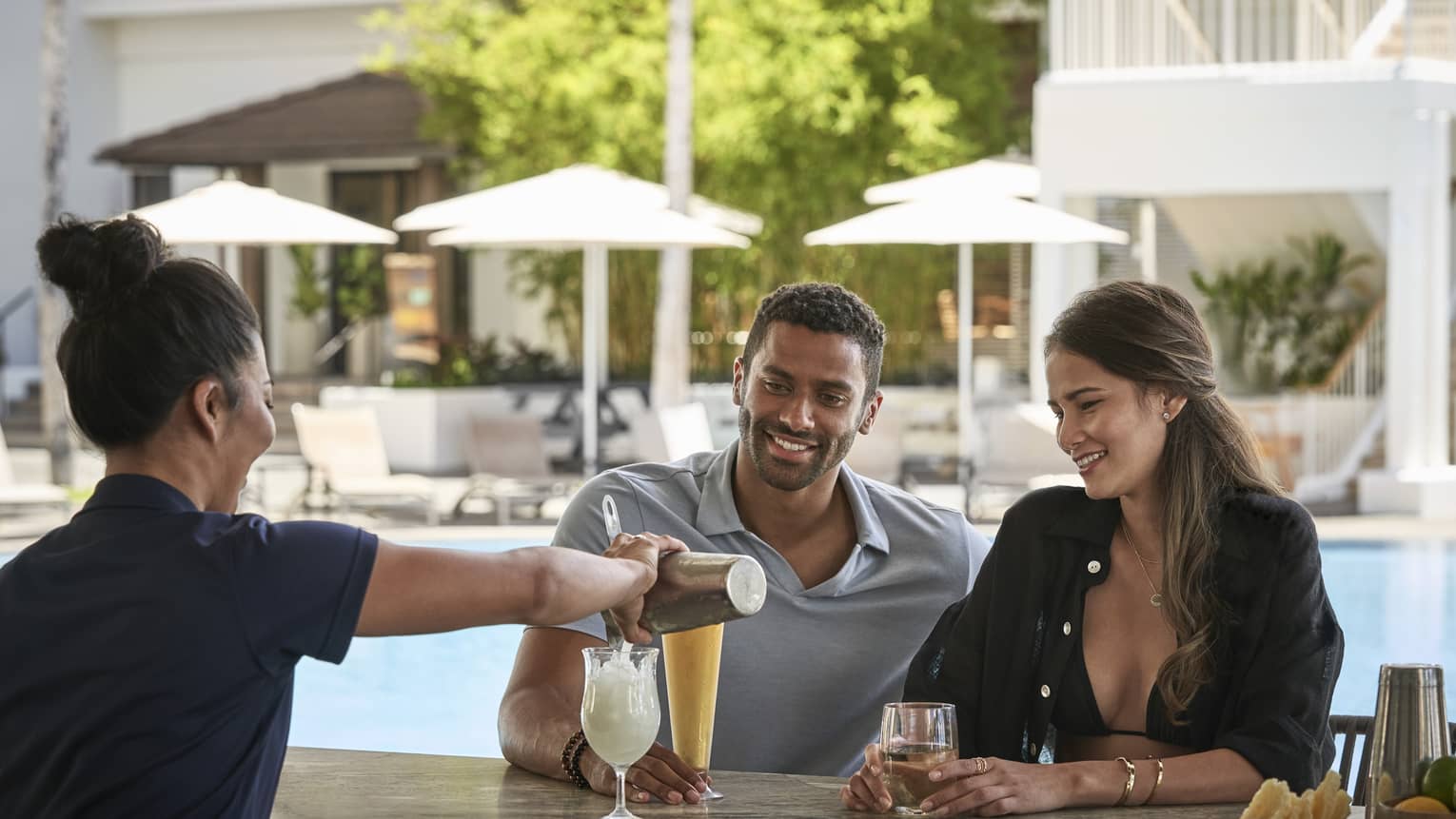Female bartender pours a tropical drink for man and woman at poolside bar