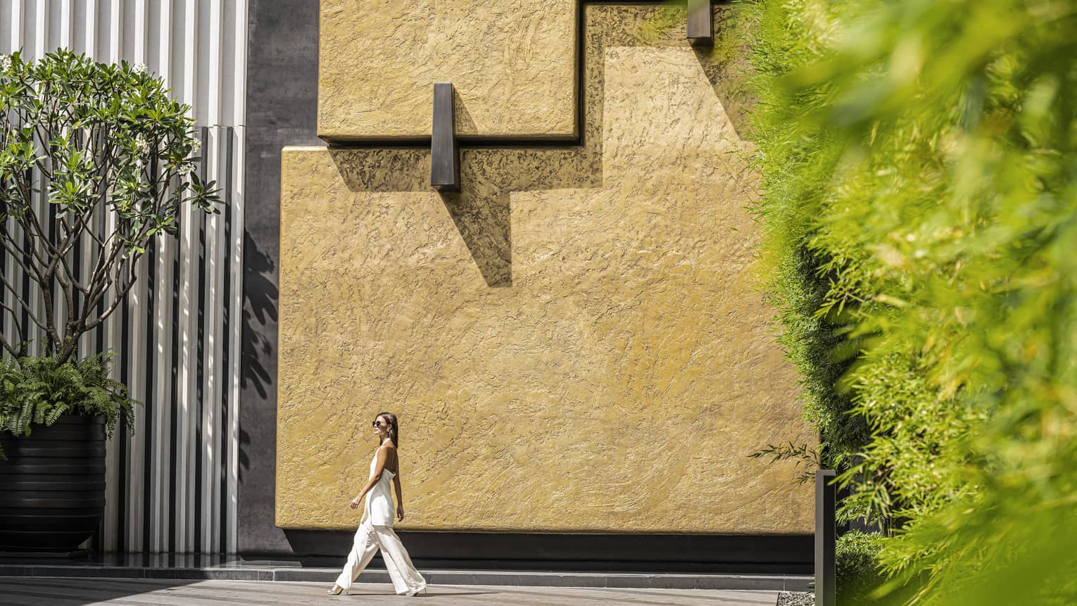 Woman walks around the exterior of the Hotel, stucco wall, oversized potted greenery