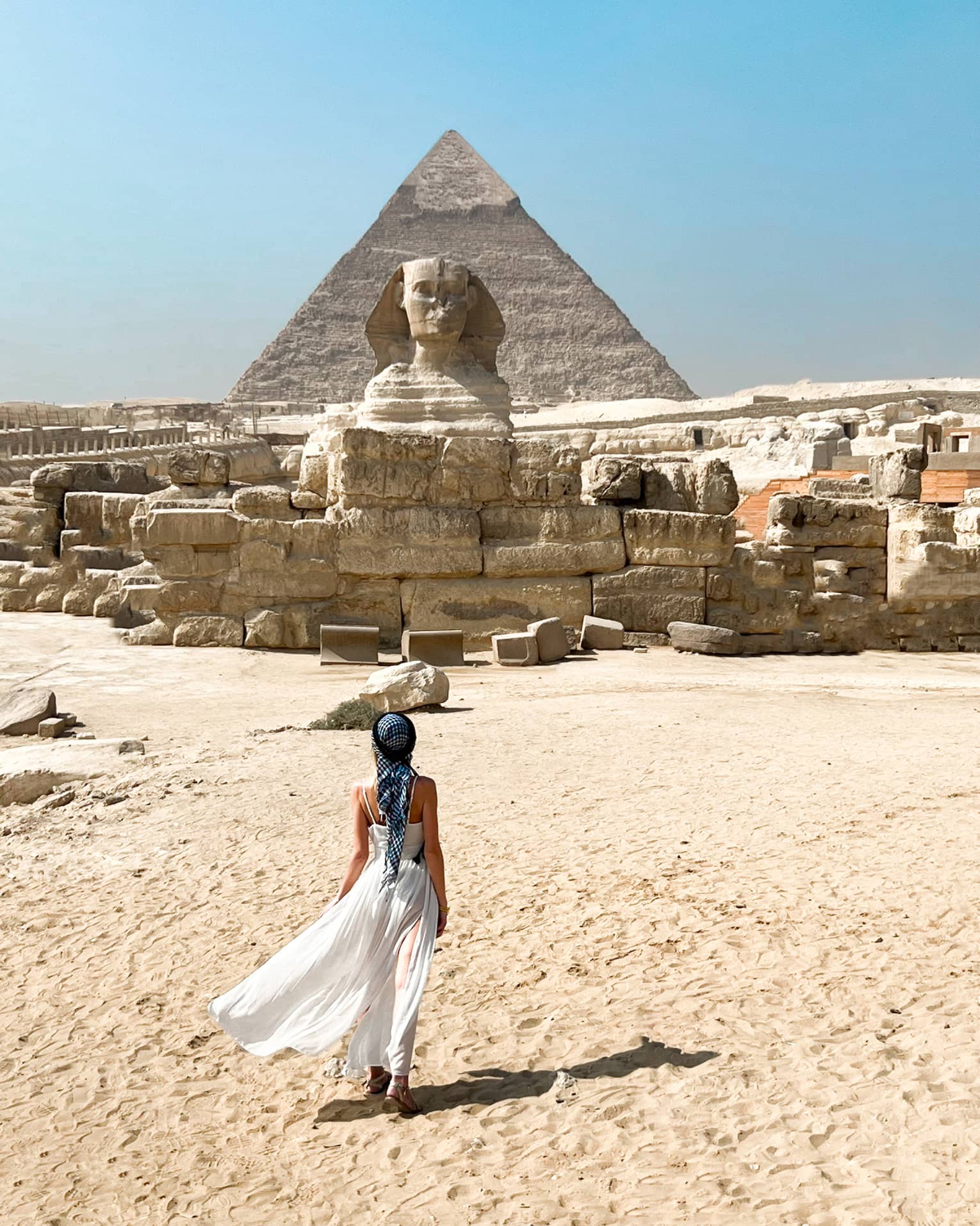 View from behind as a guest crosses golden desert sand toward the immense ruins and sphinx, a pyramid rising up beyond.