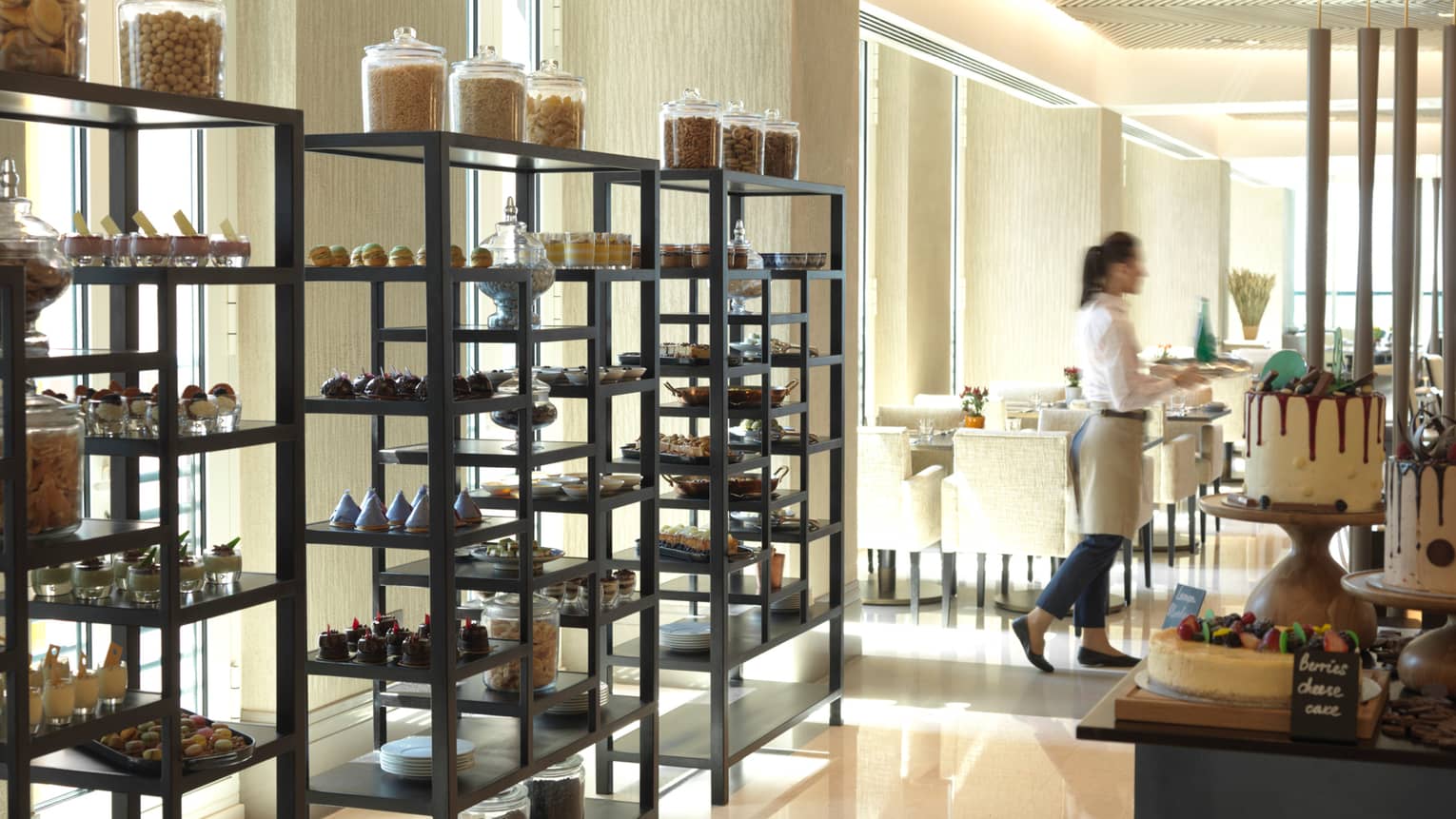 Three tall racks filled with fancy pastries and desserts in front of window, server carries tray to dining room