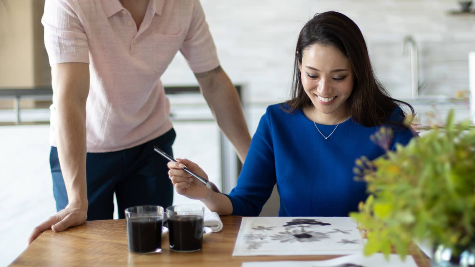 Smiling woman at table participates in Japanese ink wash painting while man stands over her should looking on