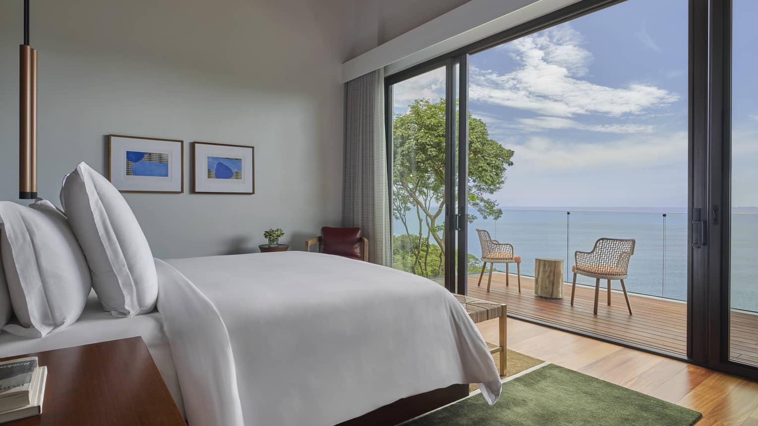 Bedroom facing wall of windows, looking out to a wooden deck with two chairs and the sea