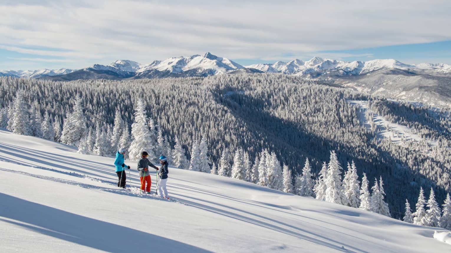 Three guests on skis pause on the ski hill, snow capped trees and mountains in distance 