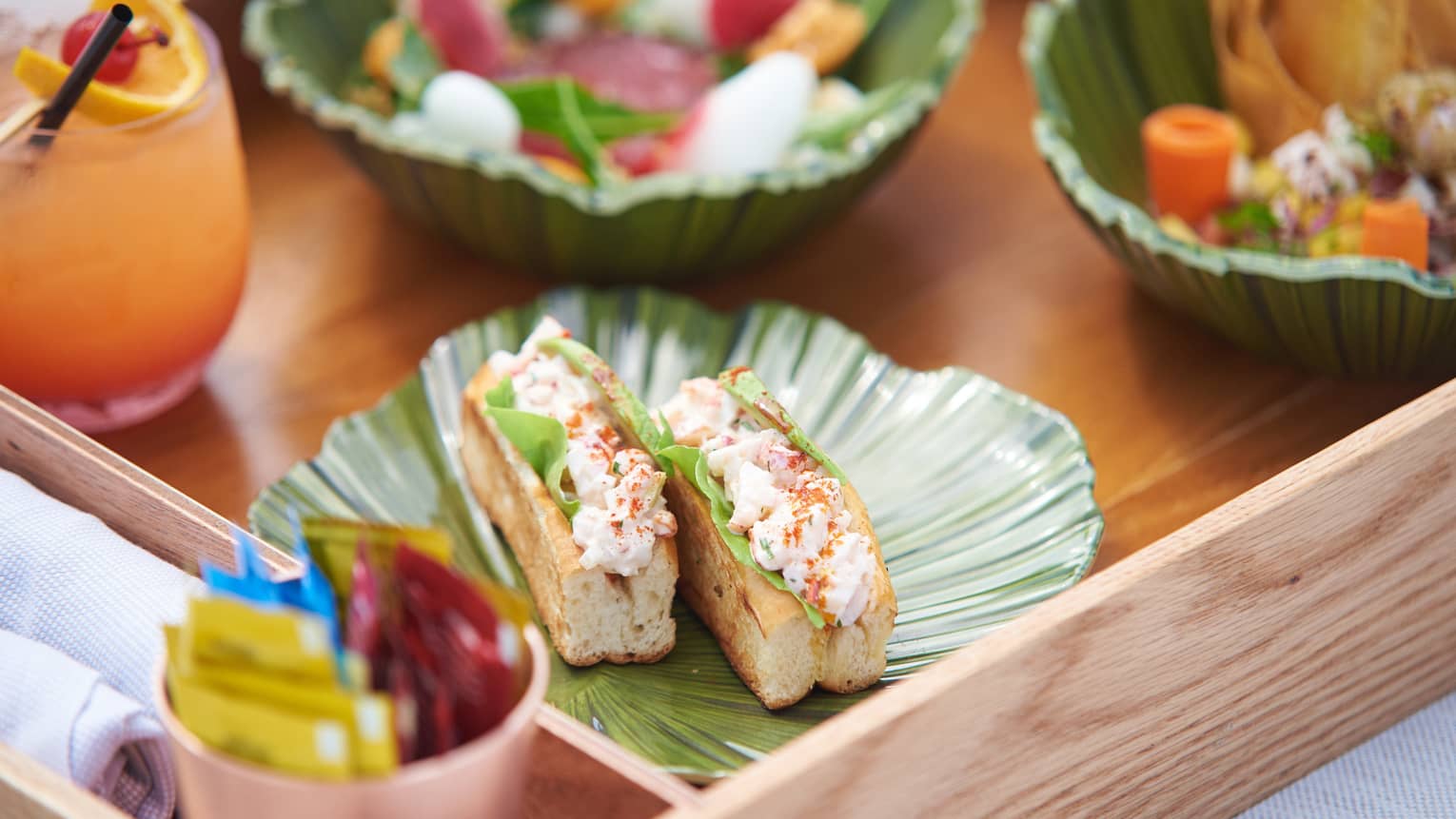 Two lobster rolls with chopped lobster meat between long buns on leaf shaped plate on tray with cocktail, salads