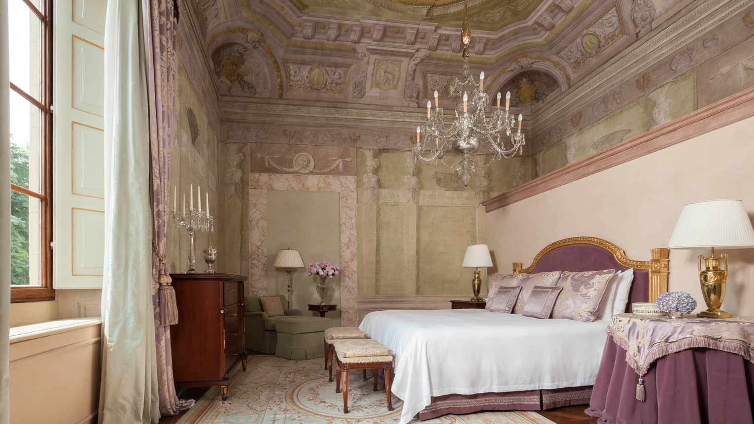 Frescoed Executive Suite bed with purple velvet headboard, nightstand with cloth under chandelier, vaulted ceiling