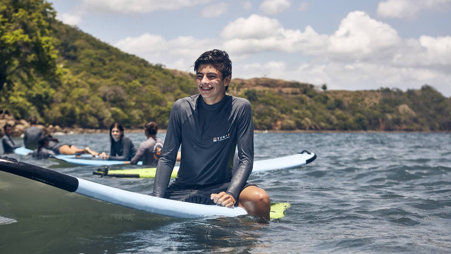 Young man with short, dark hair and wearing a dark grey long-sleeve rashguard sits on a light-blue surfboard in calm ocean waters with three other surfers sitting behind him on their boards