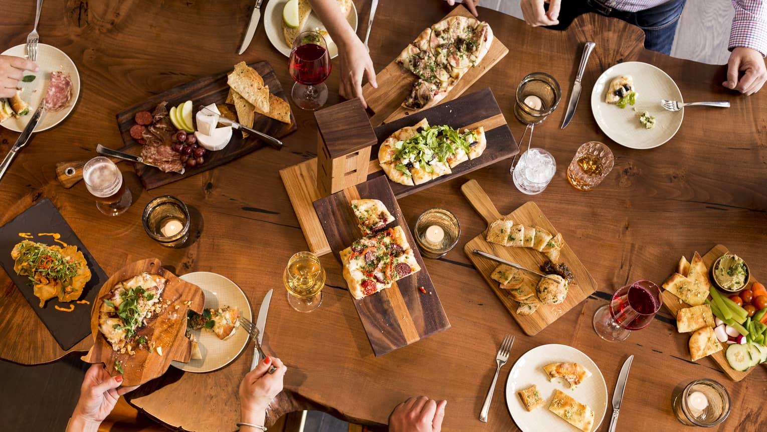 Aerial view of people dining around rustic wood table with platters of flatbreads, appetizers