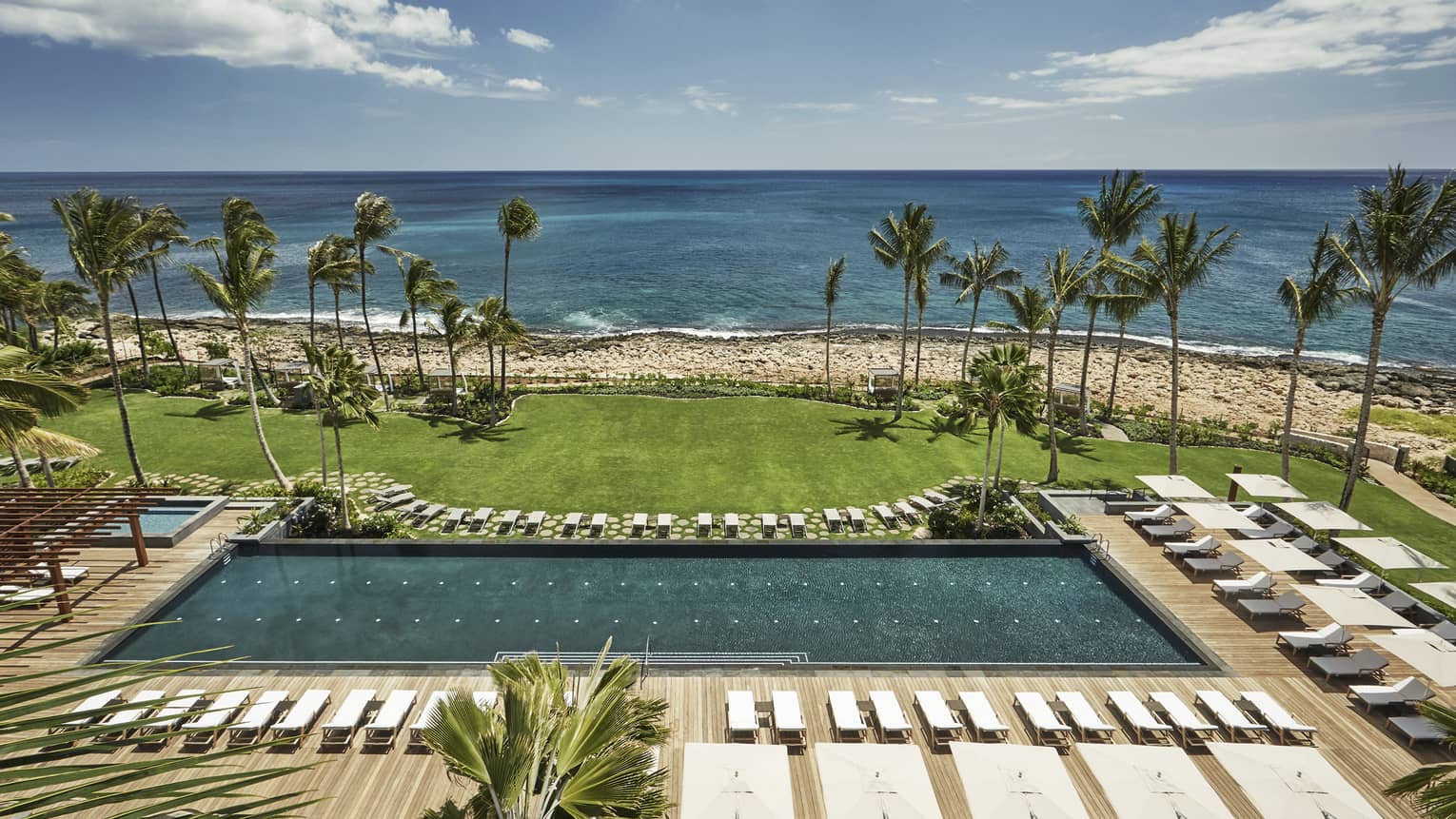 Aerial view of large outdoor swimming pool lined with white deck chairs, lawn, beach