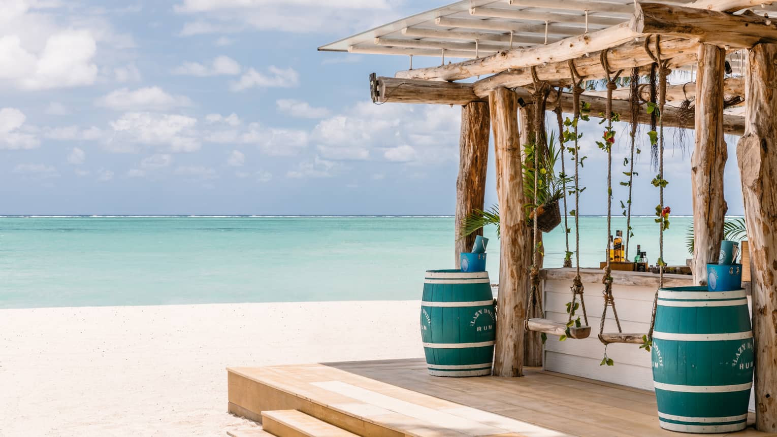 Wooden bar with pergola, two blue barrels, two swings on beach, aqua views, cloud dotted blue sky
