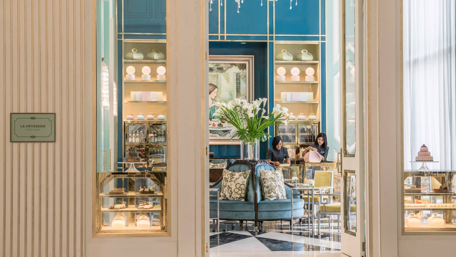 View inside La Patisserie doors with elegant blue and gold chairs, tall glass vase with white flowers, woman fastening pink box