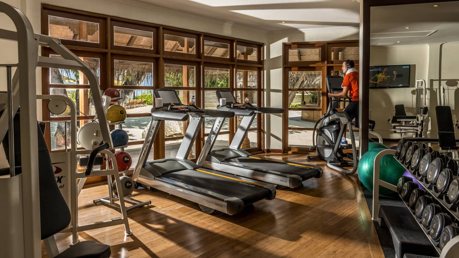 Gym with wooden floors, two treadmills, person on elliptical, free wights, windows looking out to beach