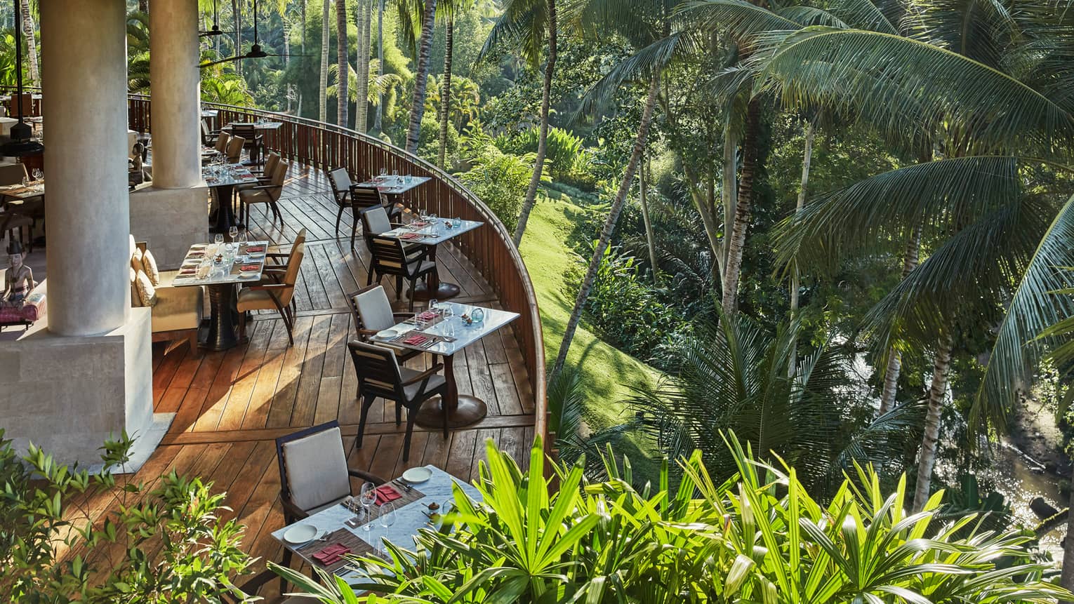 Looking down at curved wood patio of Ayung Terrace outdoor dining room, tables and chairs overlooking palm trees, forest