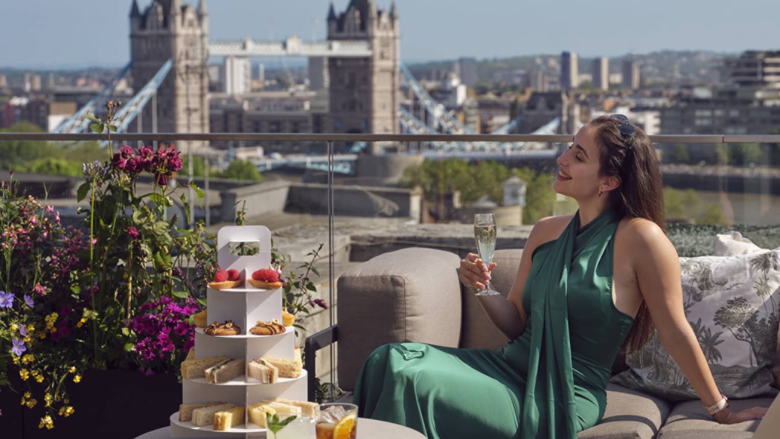 Woman in green dress holding champagne beside tea stand with confections and Tower Bridge in backdrop