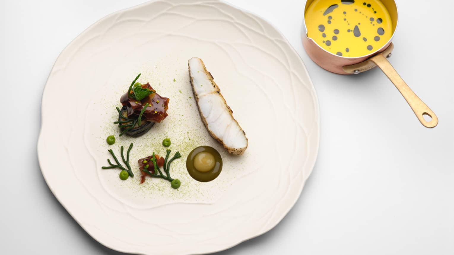 Deconstructed elegant Cornish pollock dish on ivory dish with yellow sauce in saucepot