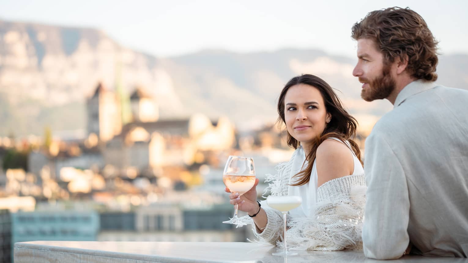 Couple leaning on terrace bar, with wine in hand, overlooking Geneva