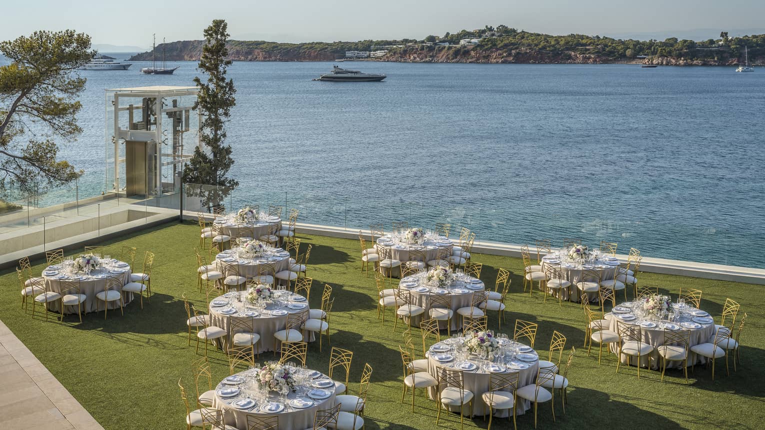 Ten round tables with gold and white chairs and flowers outside on grass overlooking ocean