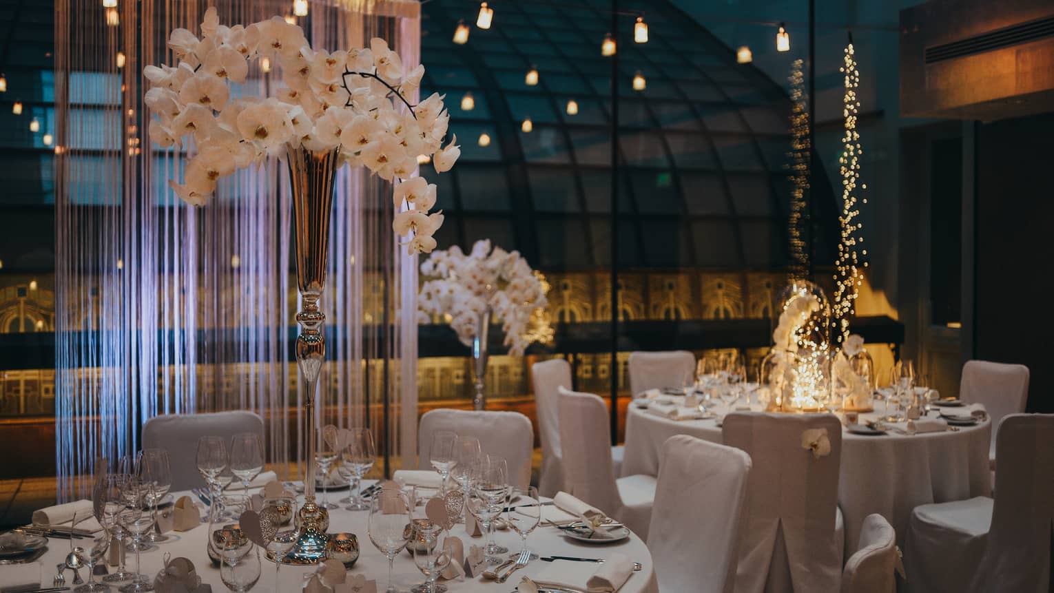 White dining tables and chairs with vase of orchids beside glass wall and reflecting lights