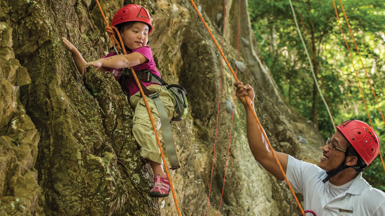 Man holds rope, helps young girl wearing helmet climb rock face