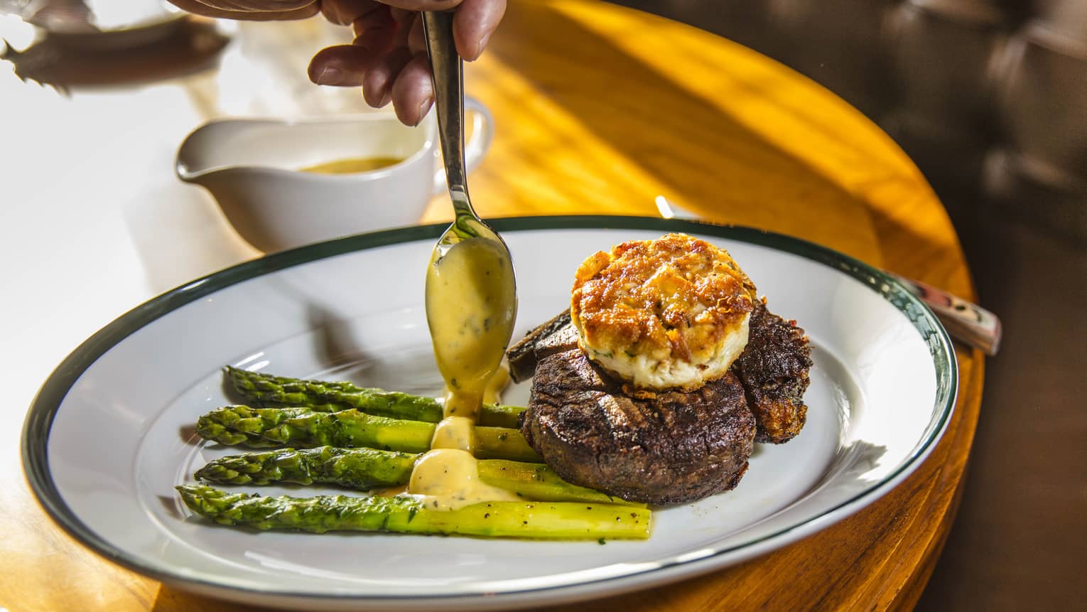 Spoon drizzles sauce over asparagus on plate with steak topped with fried cake