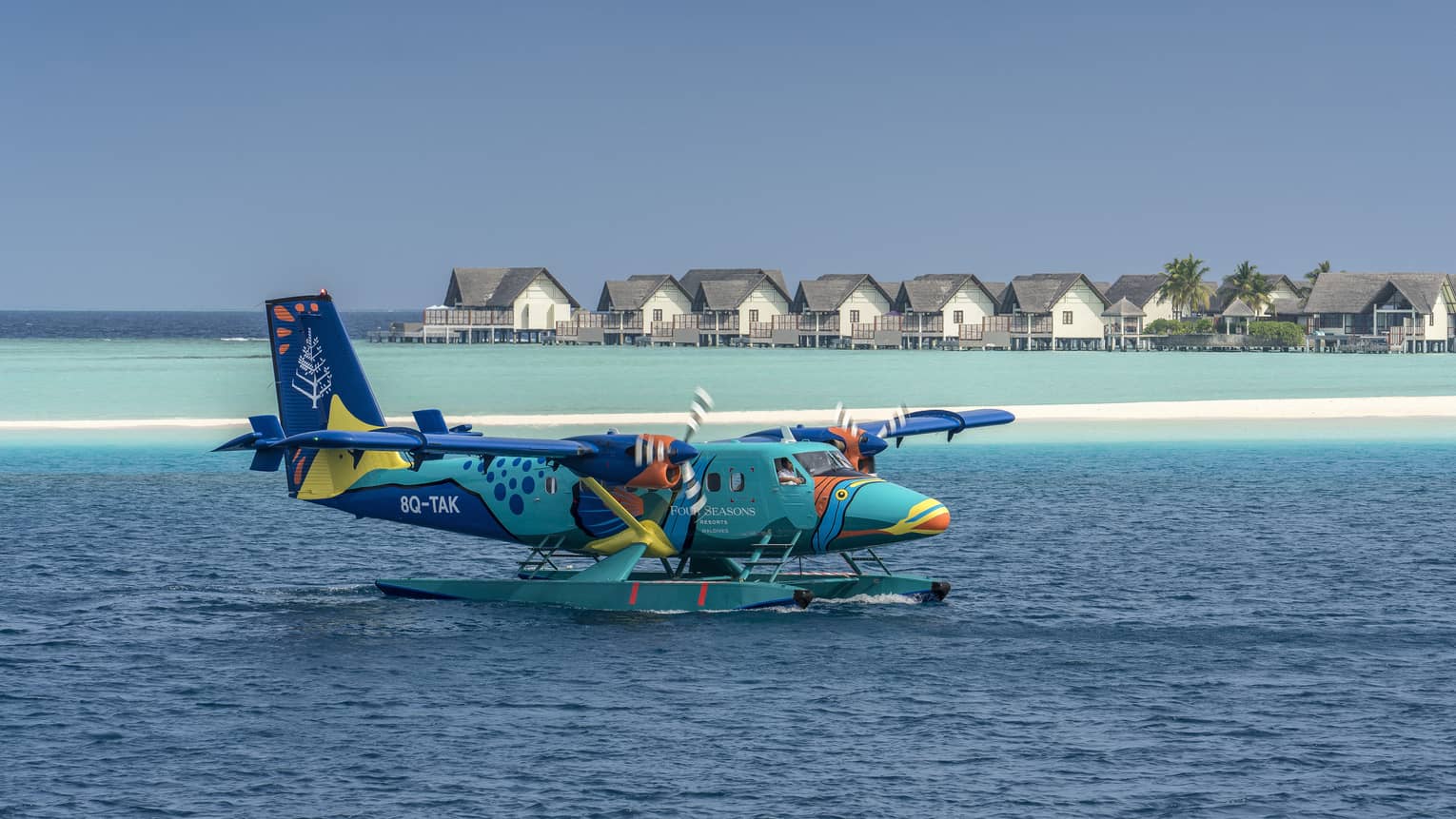 The Four Seasons sea plane landing on calm water in front of beach houses and palm trees