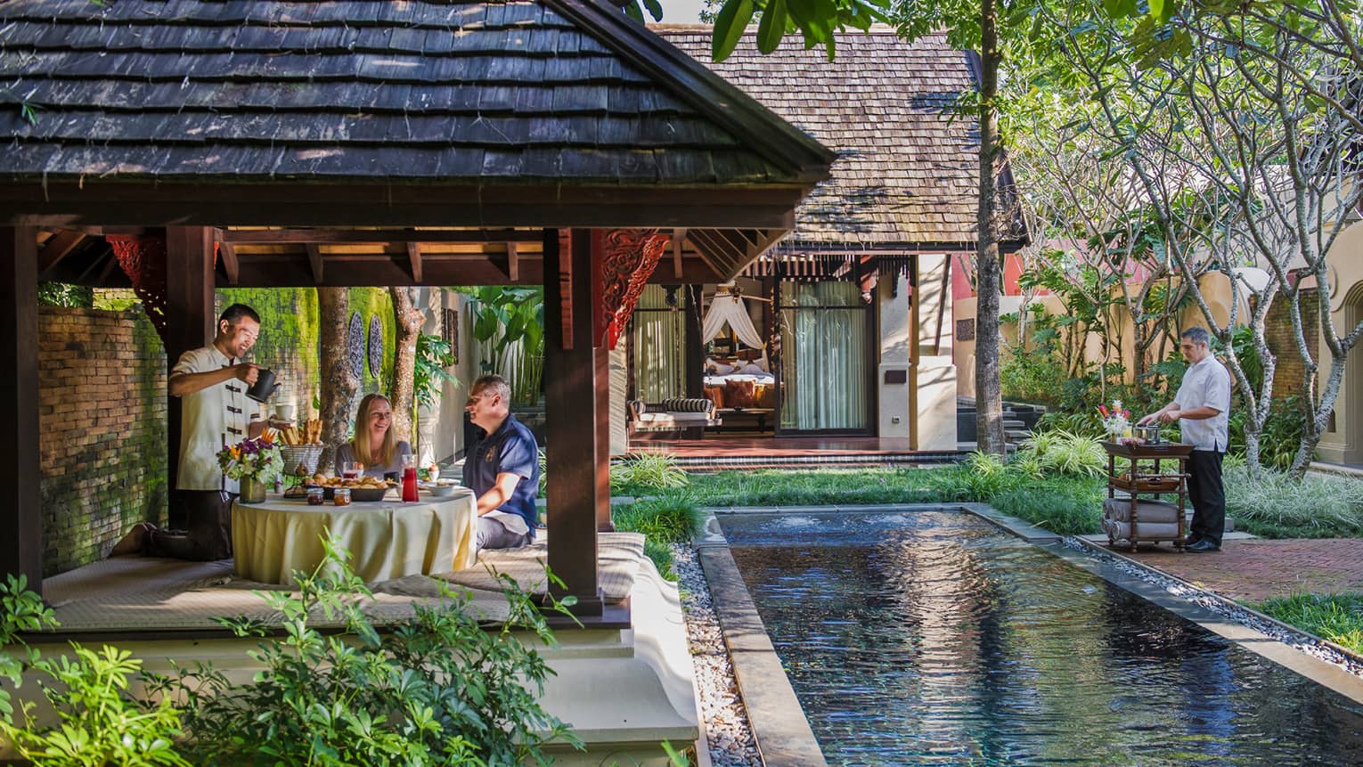 Hotel staff serve couple under dining gazebo by pond 