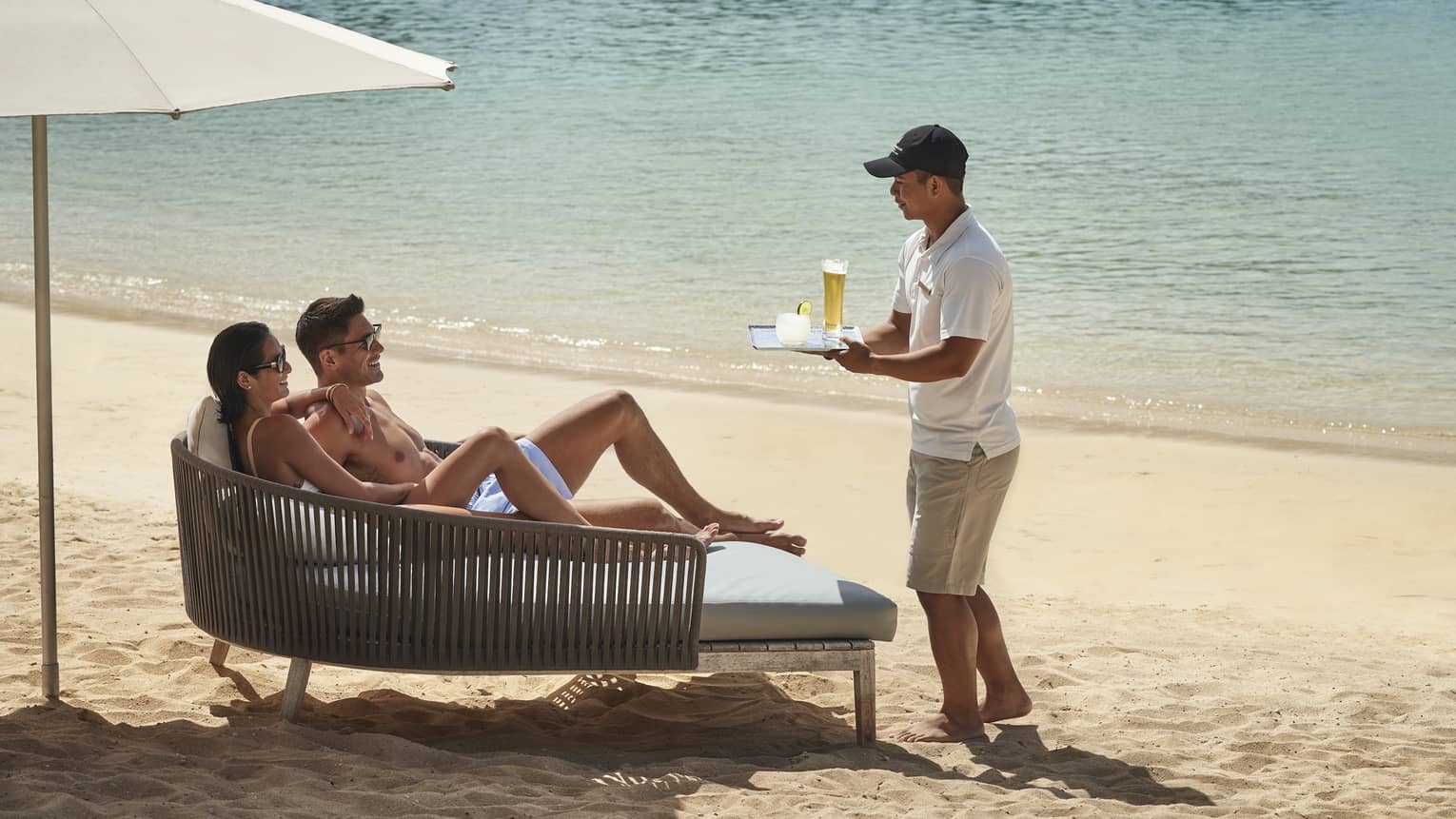 A couple lounges on a daybed on the beach while a beach attendant brings them cocktails on a tray