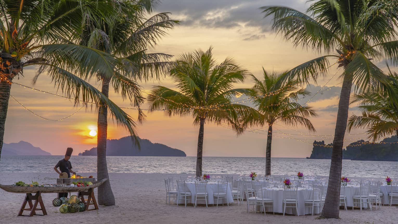 Tables set for dinner on the beach under a canopy of palm trees, a chef cooking over an open flame against the setting sun.
