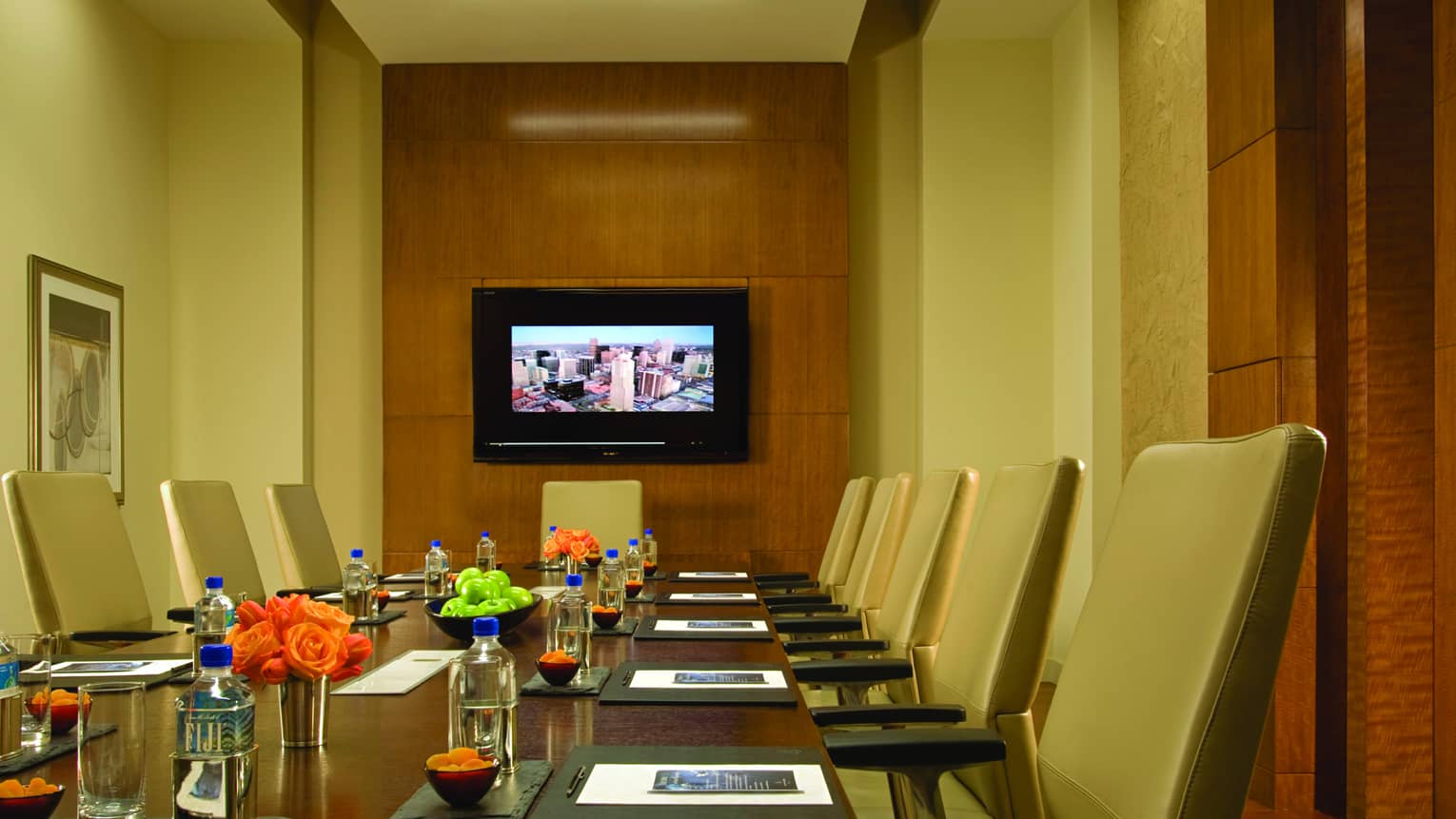 Executive boardroom table with tan leather chairs, flowers by modern wood panel wall with TV screen