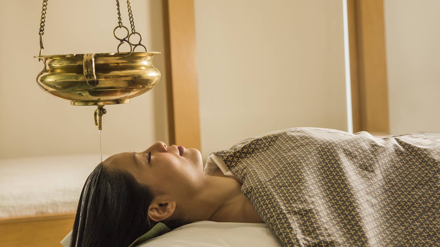 Woman lies face-up on massage table under blanket, eyes closed, gold lamp hanging above head 