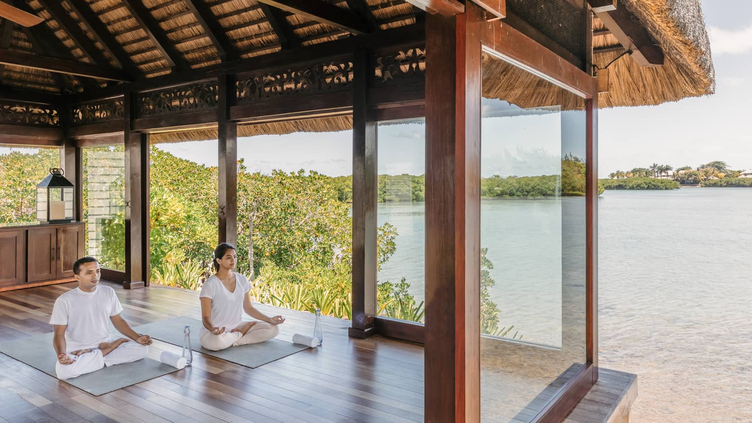 Two people wearing white seated in yoga poses in open-air spa yoga pavilion by beach