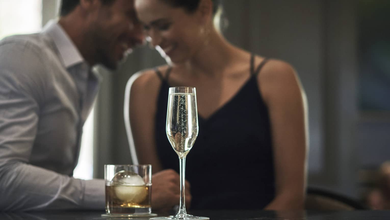 A glass of whiskey on the rocks and flute of Champagne on the bar as couple embraces in the background