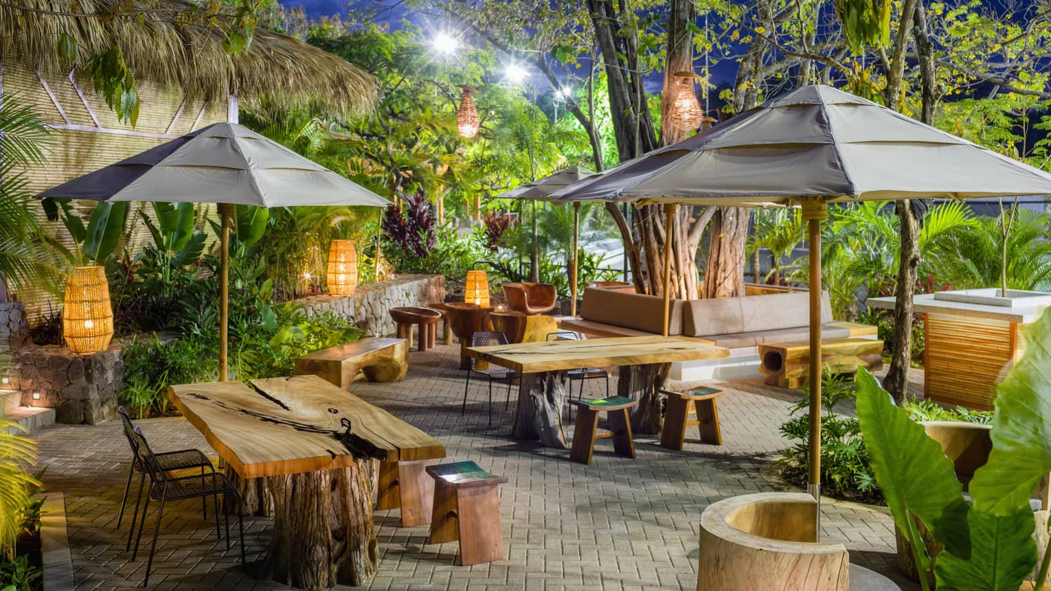 An outdoor veranda with bamboo umbrellas, wooden picnic tables, and tall leafy plants.