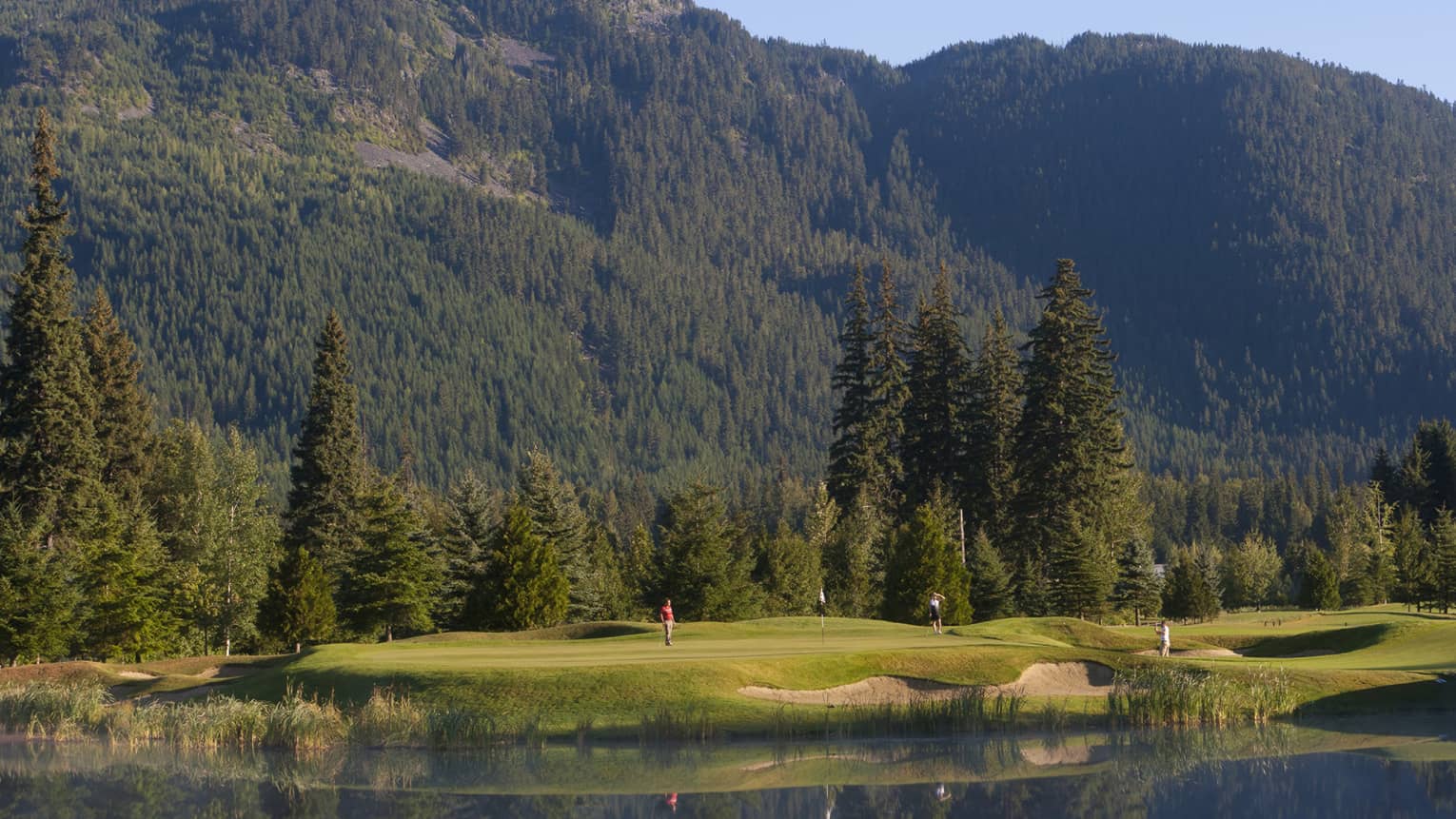 Distant golfers on a green, a pond in front, evergreens behind, dwarfed by a treed mountain filling most of the frame beyond.