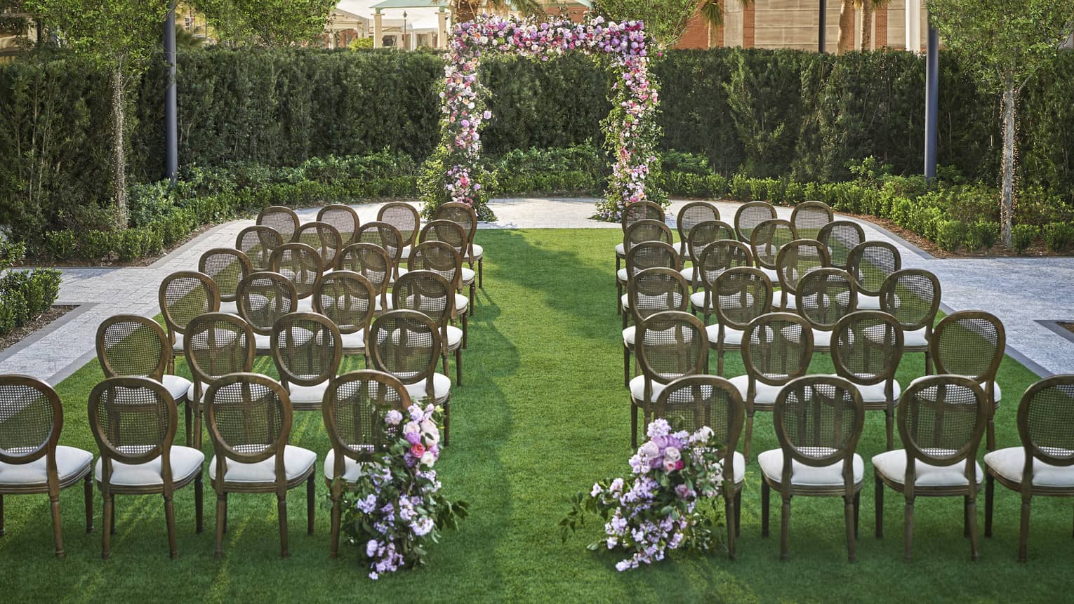 Lawn set up for wedding ceremony with rows of chairs, flower arch and rounded hedges