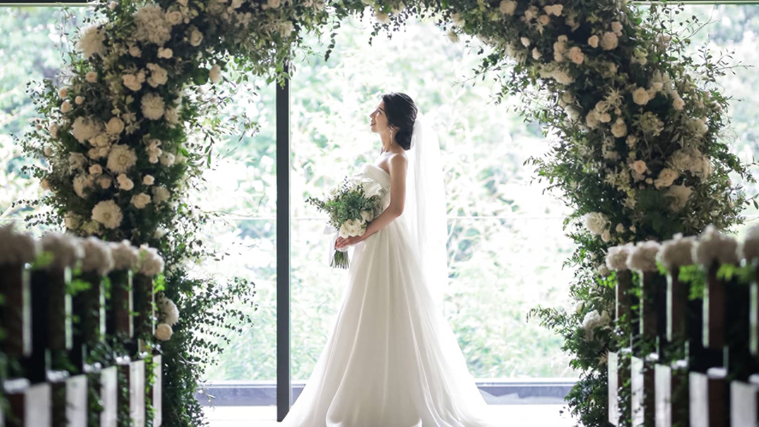 Bride stands under round wedding altar covered in white flowers by sunny window
