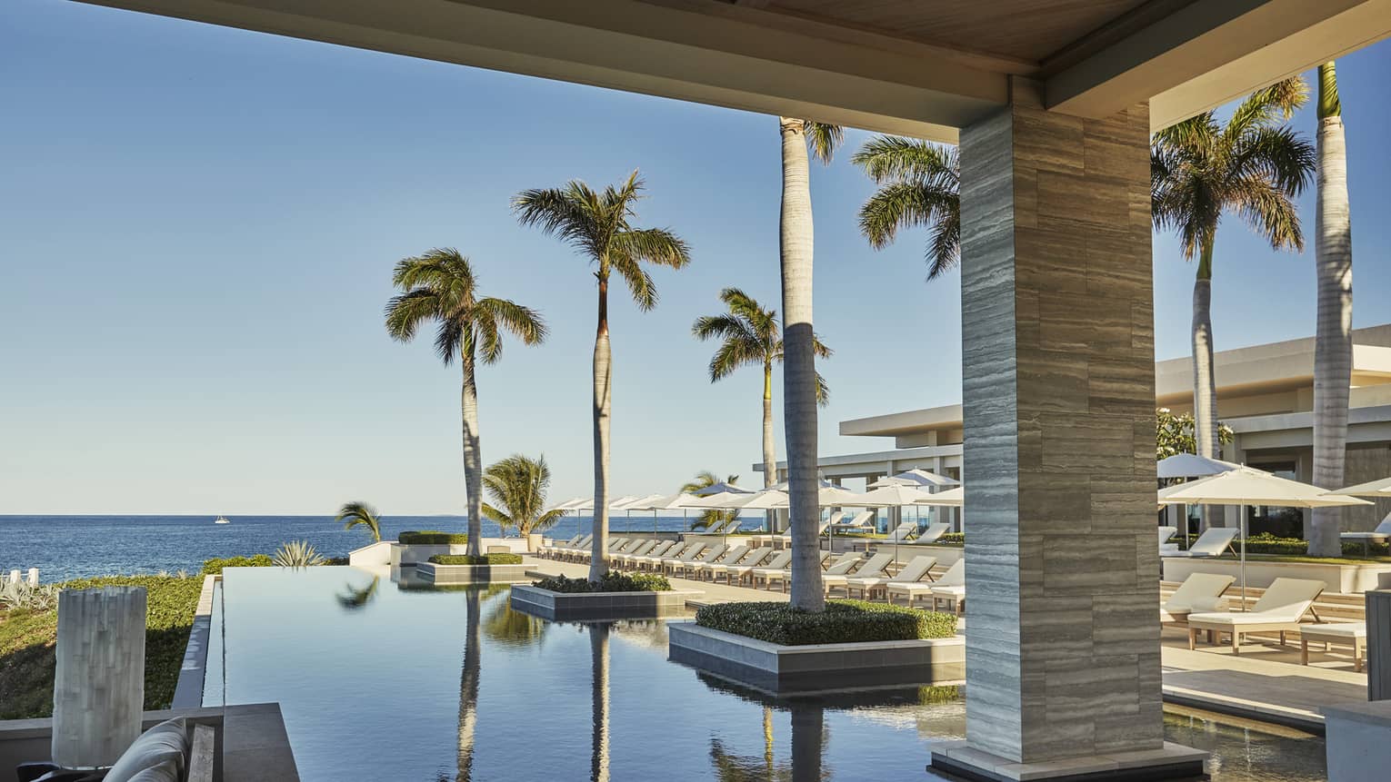 Infinity pool with potted palm trees next to row of white lounge chairs, umbrellas, ocean view